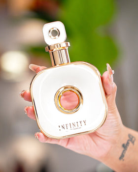 A beautifully manicured hand holding the luxurious Armaf Infinity Gold perfume bottle. The bottle is uniquely designed in glossy white and gold, featuring a hollow center, sophisticated branding, and an elegant, eye-catching shape. Soft greenery and subtl
